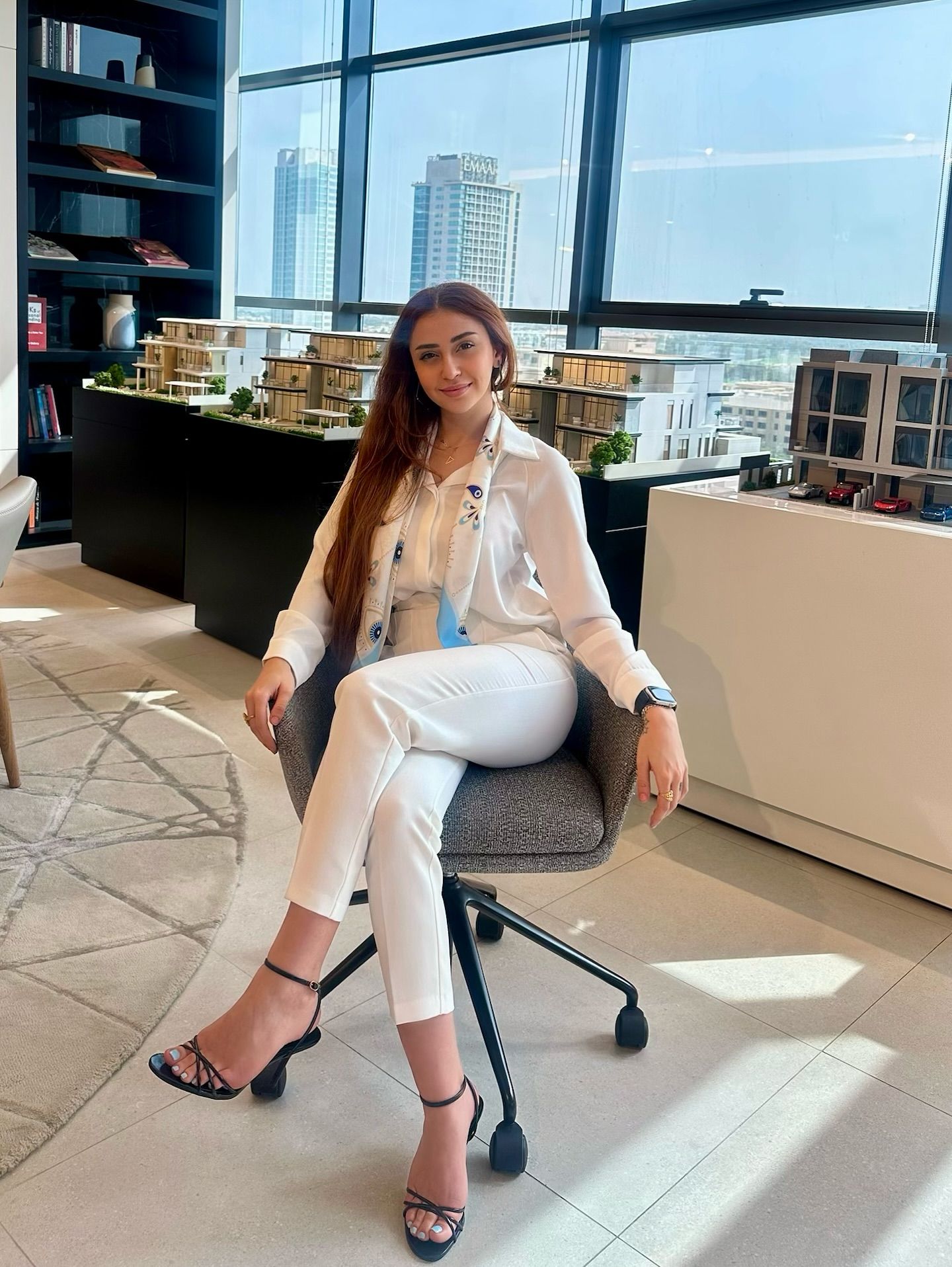 Image of Hayat Nurdeen | حياة نورالدين from Instagram white outfit, long-sleeve shirt, white pants, scarf, black high-heeled sandals, watch, office, modern background, barefoot