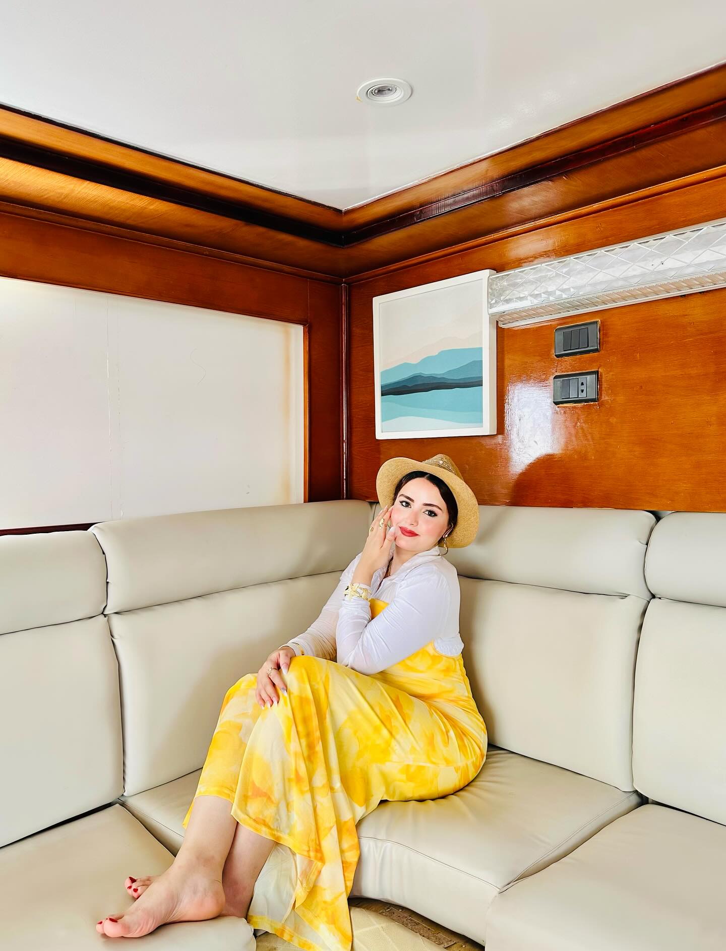Image of AYA Elboghdady from Instagram yellow dress, white top, straw hat, barefoot, bracelet, ring, beige sofa, wood paneling, artwork