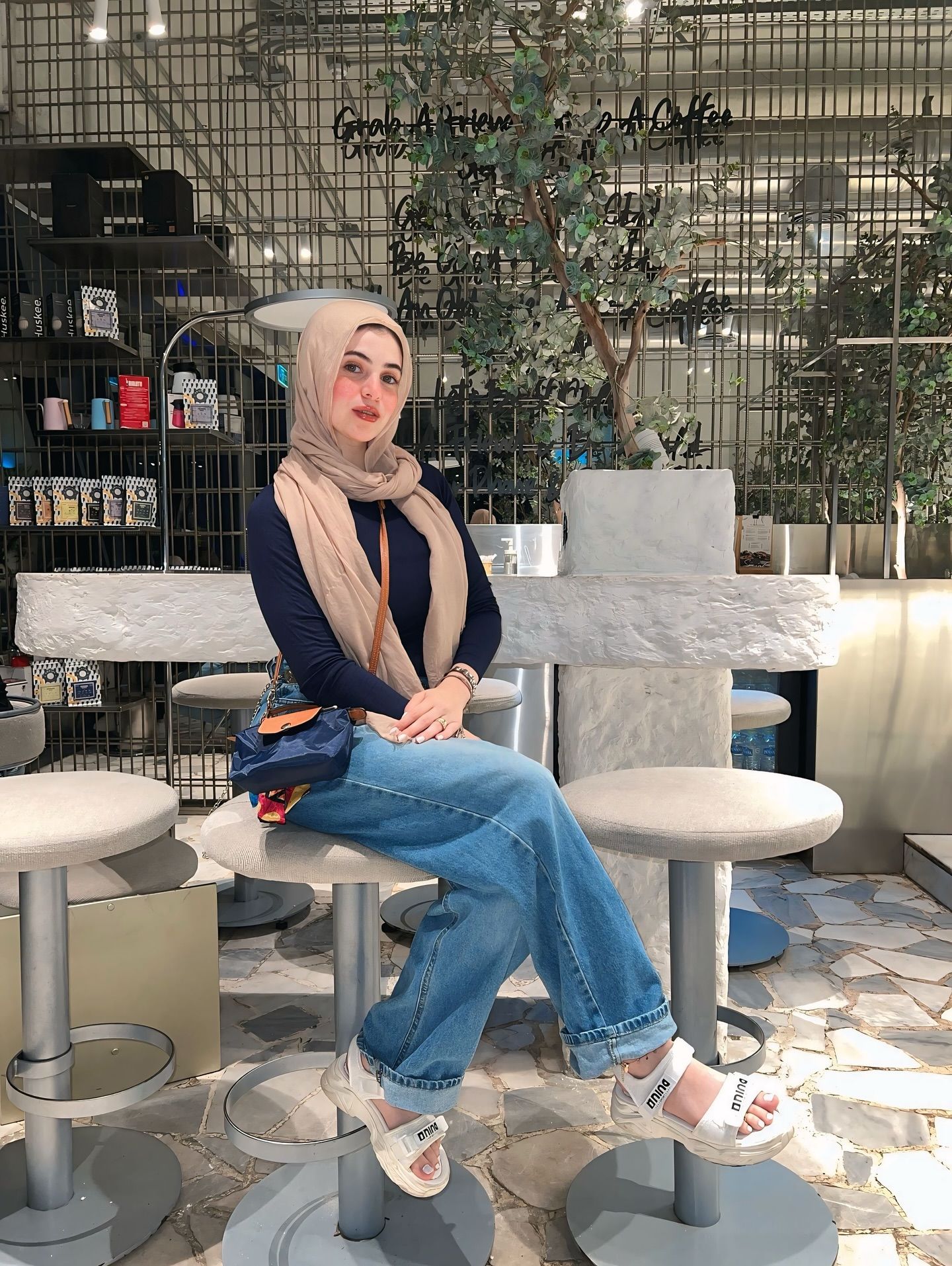 Image of Rehab Amgad from Instagram beige scarf, navy top, blue jeans, slide sandals, crossbody bag, indoor, cafeteria, trendy