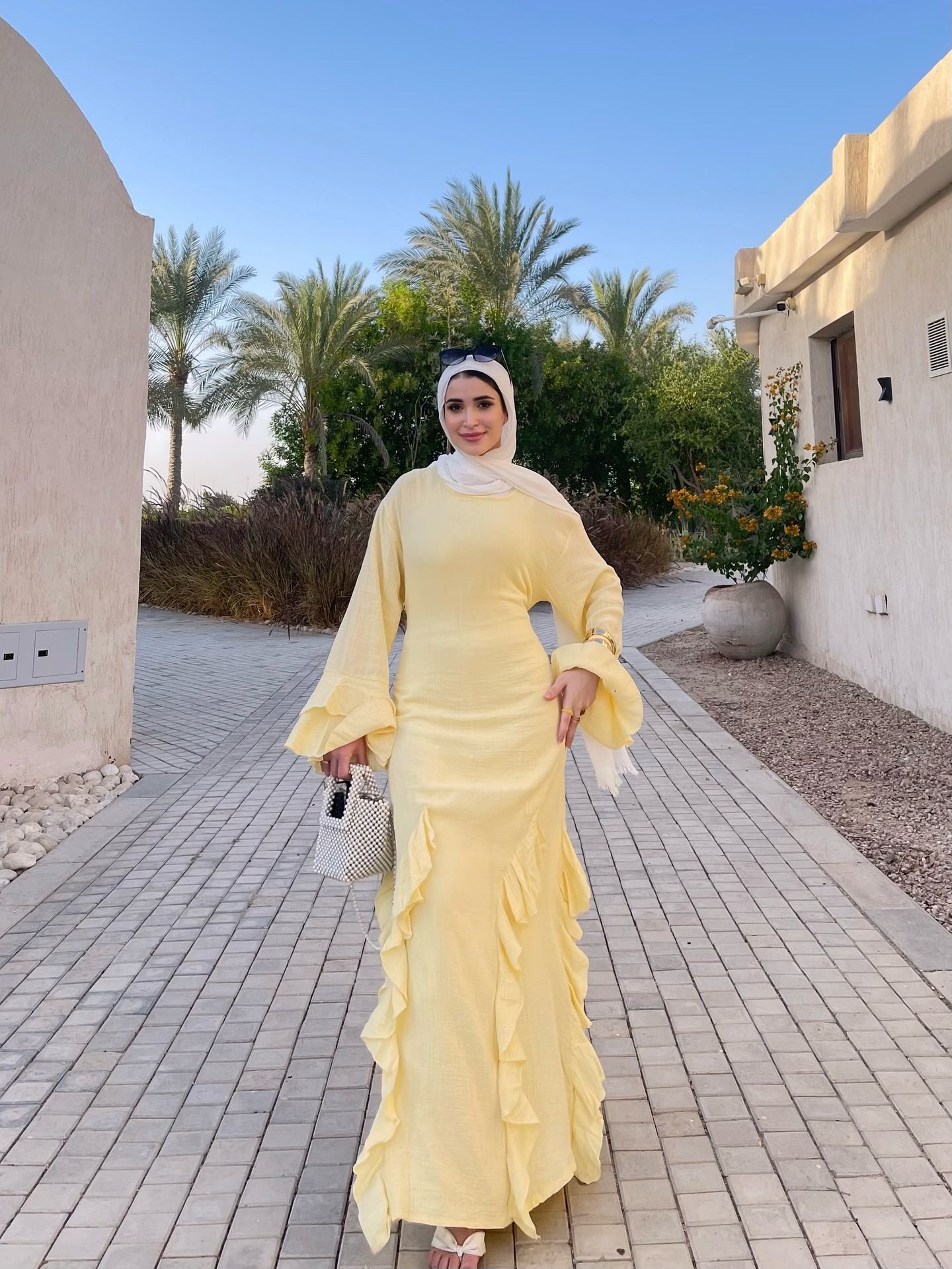 Image of 🅝🅐🅓🅐 🅖🅐🅜🅐🅛 🅨🅐🅗🅘🅐 from Instagram yellow dress, long sleeves, ruffles, white hijab, sunglasses, beaded bag, block heels, summer fashion, outdoor background