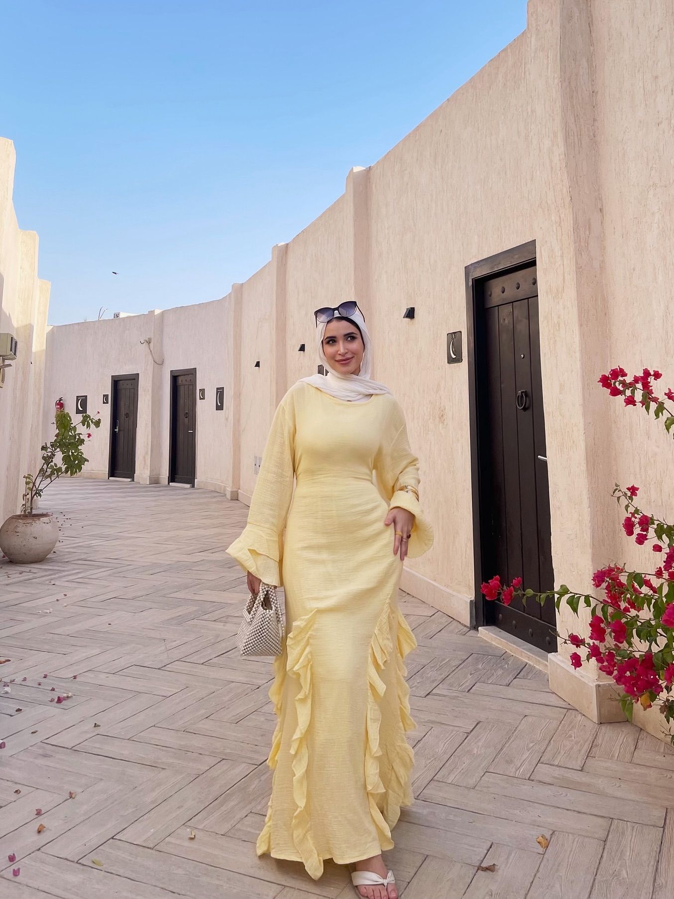 Image of 🅝🅐🅓🅐 🅖🅐🅜🅐🅛 🅨🅐🅗🅘🅐 from Instagram yellow dress, ruffled design, white hijab, white sandals, woven handbag, sunglasses, outdoor setting, arched alleyway, floral decor, barefoot