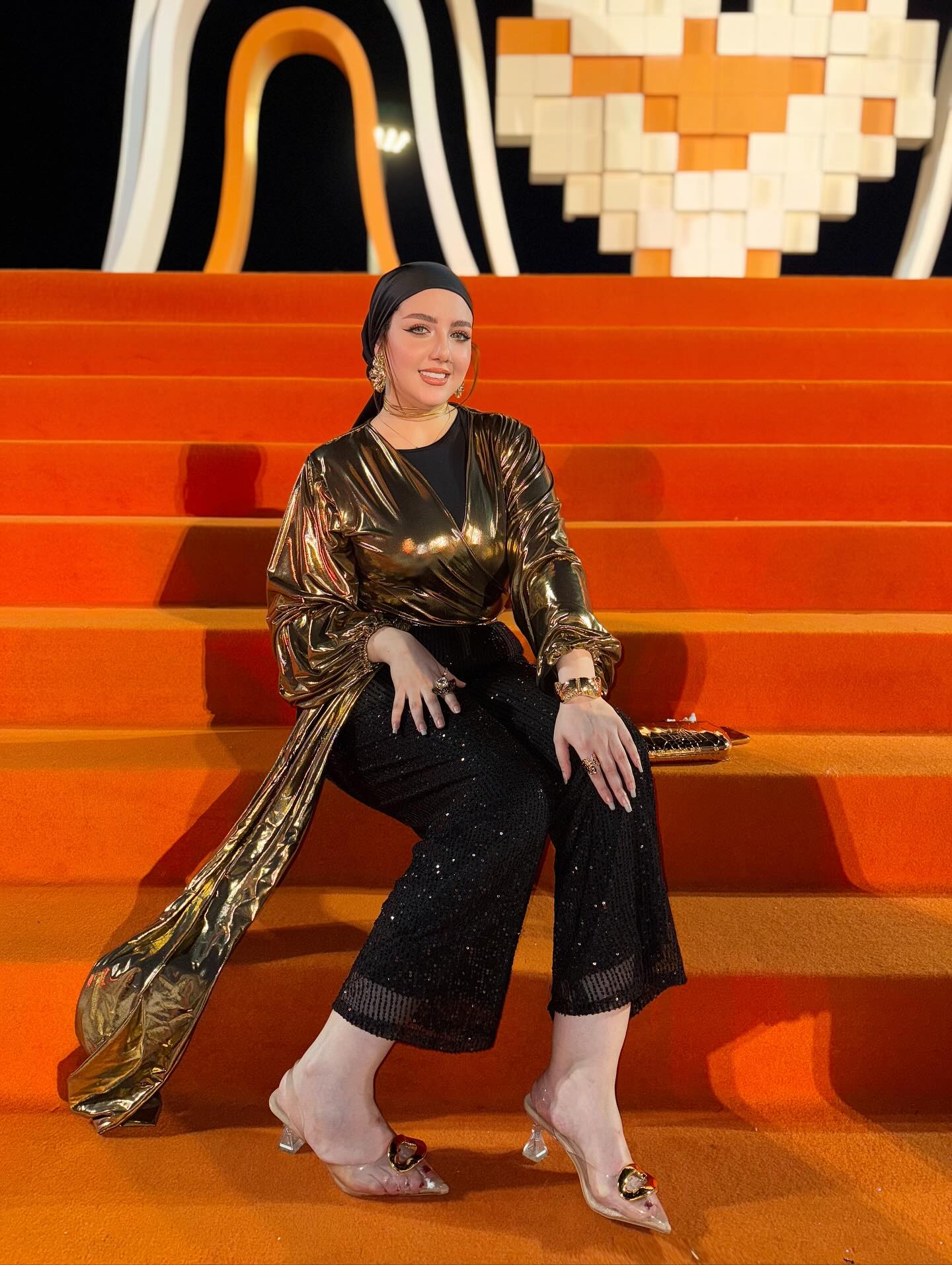 Image of makeup by merna t3ima from Instagram metallic wrap top, black sequin pants, transparent heels, gold accessories, black headscarf, gold clutch, orange stairs, modern background