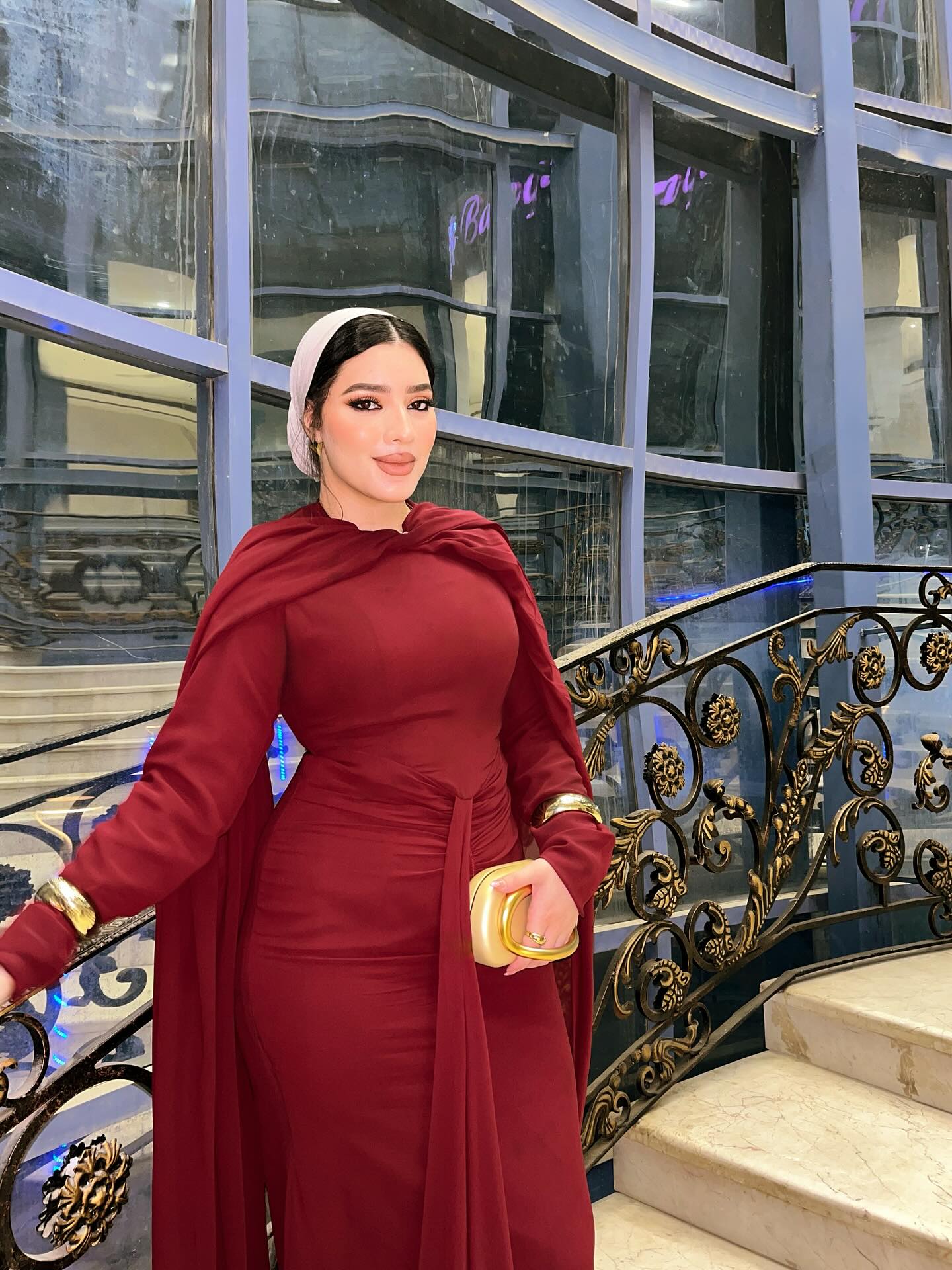 Image of SHERRY MOHAMED | شيري محمد📍 on Instagram: "صور من السنه اللي فاتت بس جه وقتها 😂💗" from Instagram maroon gown, long sleeves, floor-length dress, drape, headscarf, bracelets, gold clutch, elegant, staircase, ornate railing