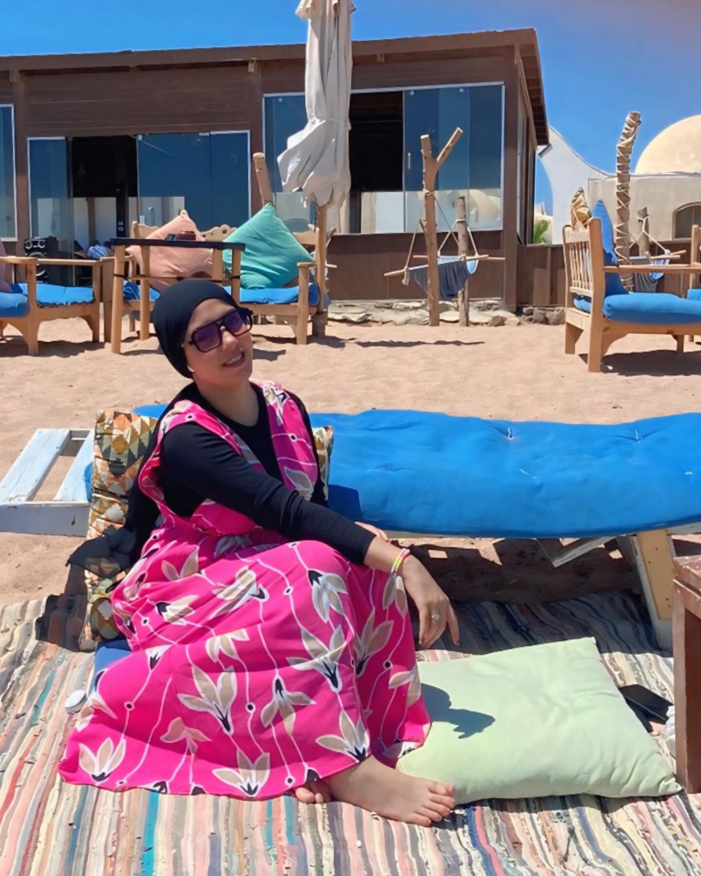 Image of NaDa NabiL-ندي نبيل from Instagram bright pink dress, floral pattern, black top, sunglasses, headscarf, bracelets, barefoot, beach setting, outdoor seating