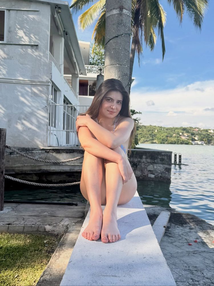 Woman in bikini top sitting barefoot on a dock by a lake.