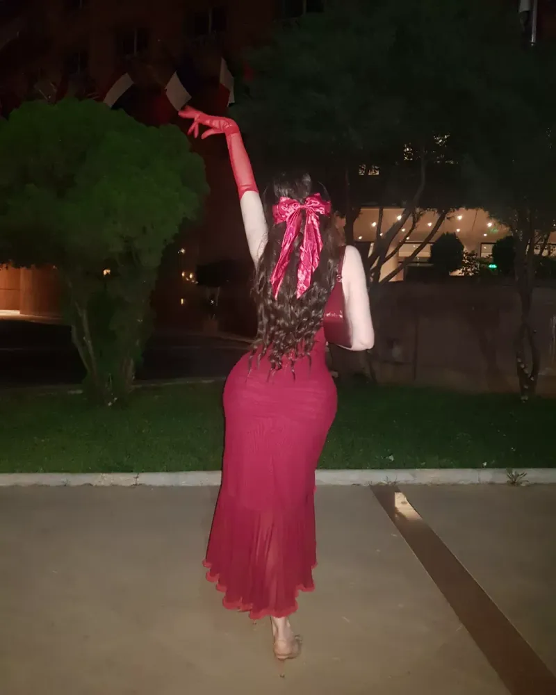 Woman in a fitted red dress with gloves, barefoot in an outdoor evening setting.
