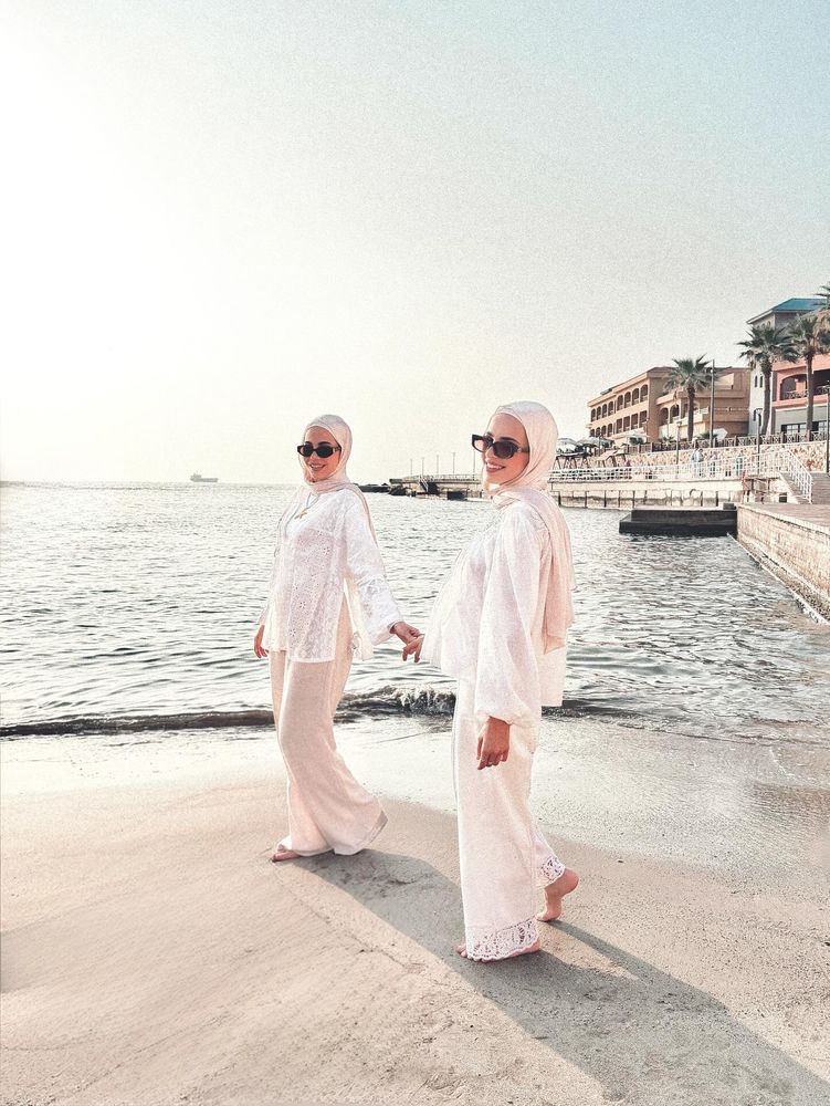 Two women in white outfits and sunglasses walking on the beach.
