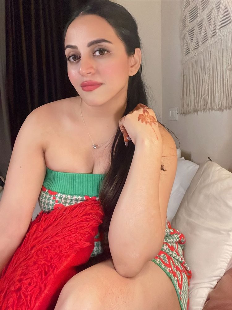 Woman in a colorful strapless dress with henna on her hand, sitting on a bed.