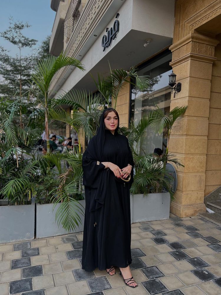 Girl in black abaya and strappy sandals among greenery.