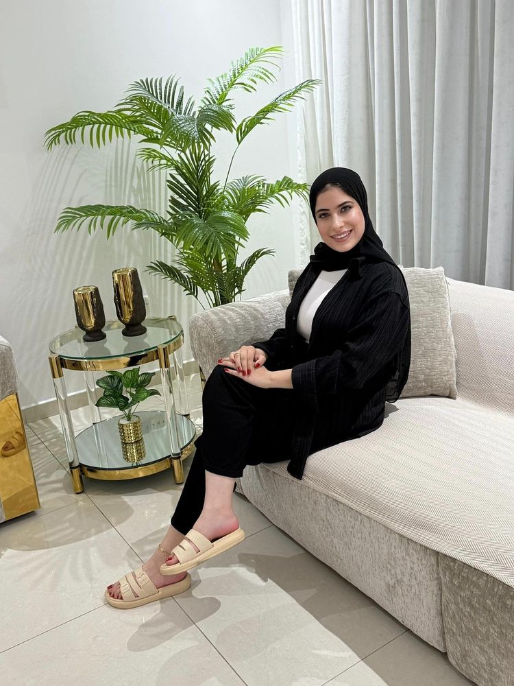 Young woman in black oversized shirt and beige slide sandals sitting on a couch.