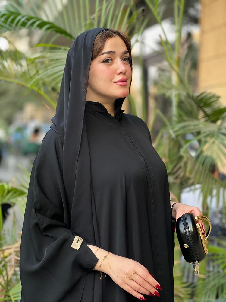 Woman in black abaya and matching hijab holding a handbag in a garden setting.
