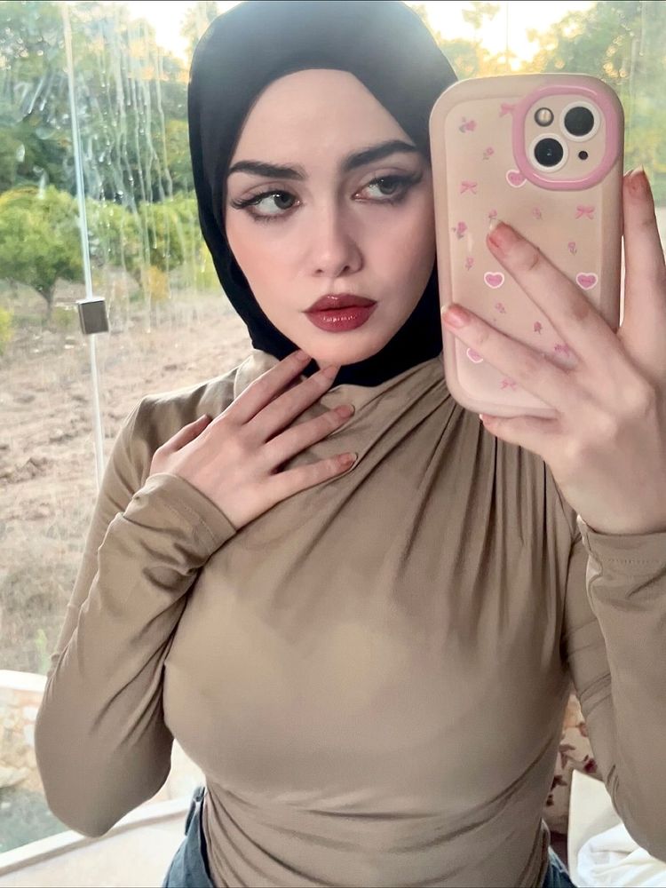 Woman in taupe long-sleeve top and black hijab, posing in a reflective setting.