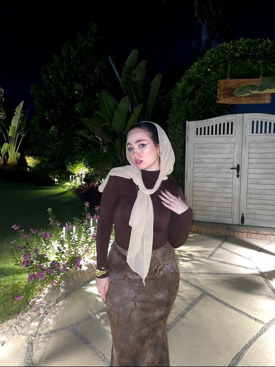 Ashrkat wearing a brown top and patterned skirt with a hijab in a garden setting.