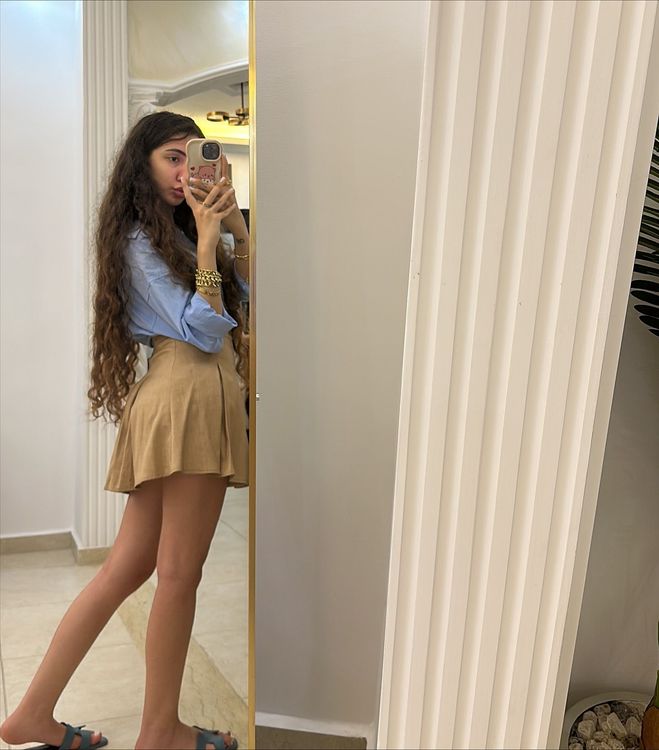 Girl in light blue button-up shirt and tan mini skirt with stylish slides in a bright setting.