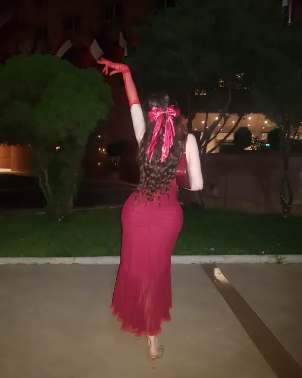 Woman in a fitted red dress with gloves, barefoot in an outdoor evening setting.
