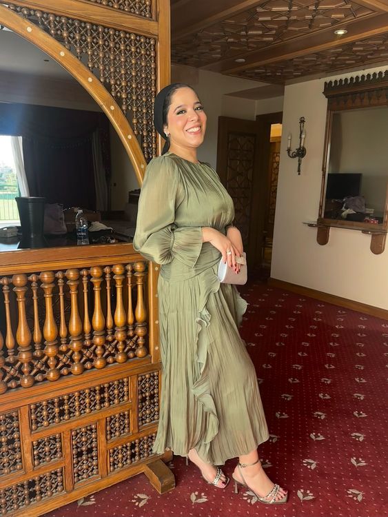 Woman in olive-green pleated dress and open-toe heels standing beside intricate wooden decor.