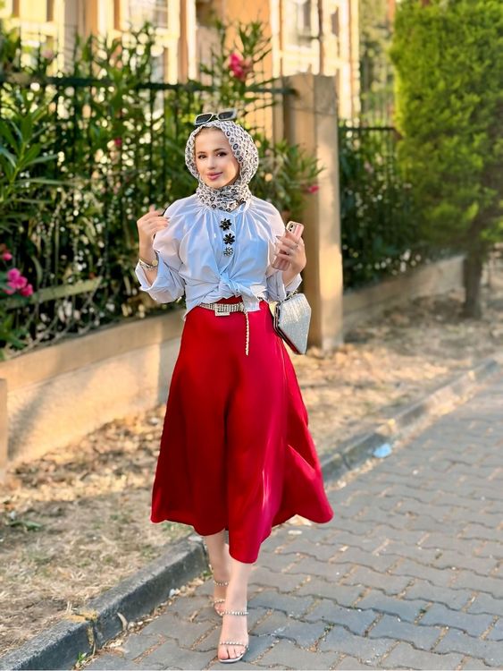 Woman in pastel blouse and red midi skirt with strappy sandals in a garden setting.