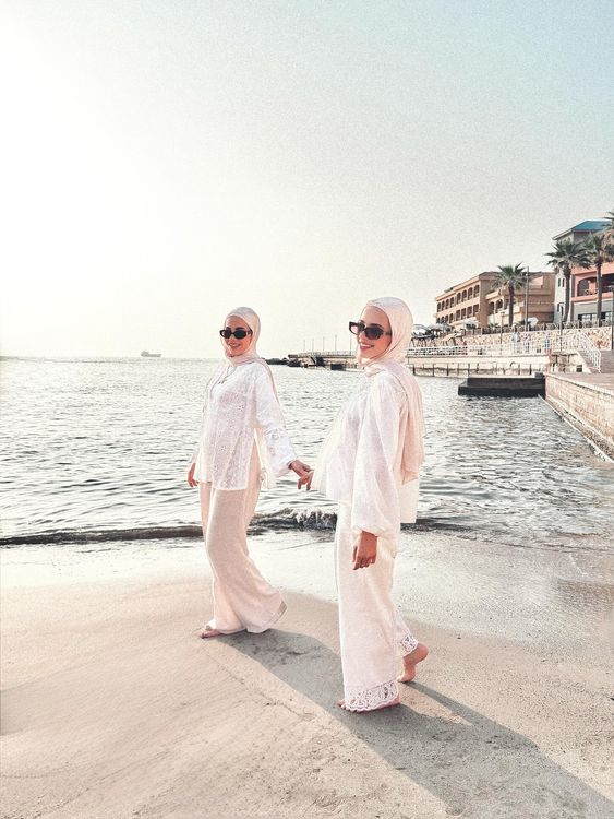 Two women in white outfits and sunglasses walking on the beach.