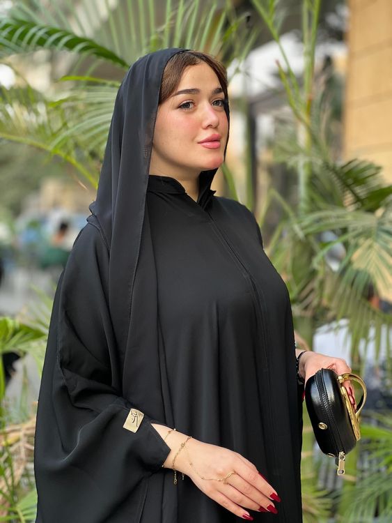 Woman in black abaya and matching hijab holding a handbag in a garden setting.