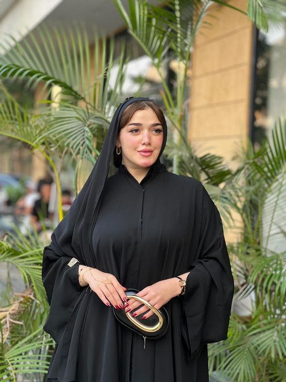 Woman in a black abaya and handbag with green plants in the background.