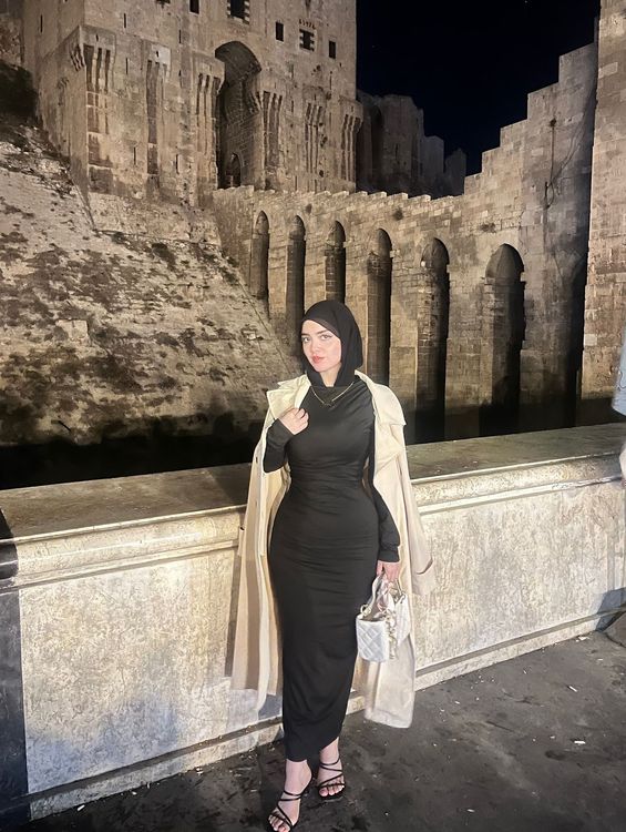 Woman in a black dress and strappy sandals against a historic stone backdrop.