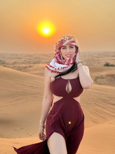 Woman in burgundy cut-out dress and hijab against a desert sunset.