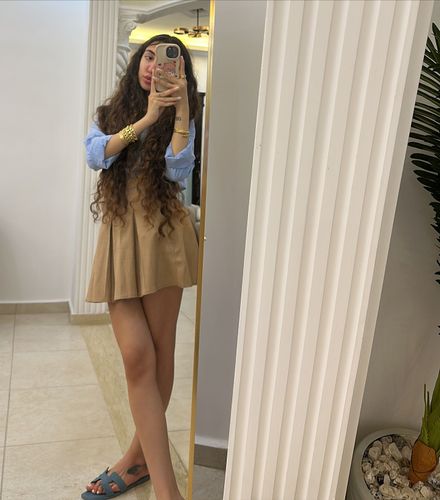 Girl in light blue top and beige mini skirt wearing blue slides in a bright indoor setting.