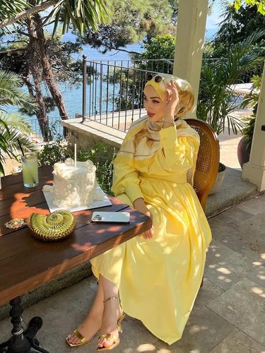 Woman in yellow dress and gold sandals sitting by the water with cake.