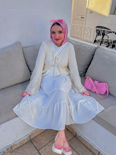 Woman in a white dress and elegant sandals with pink accessories and hijab sitting on a sofa.