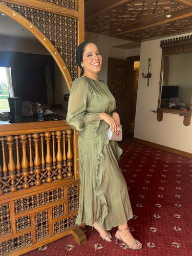Woman in olive-green pleated dress and open-toe heels standing beside intricate wooden decor.