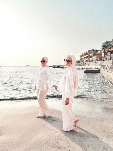 Two women in white outfits and sunglasses walking on the beach.
