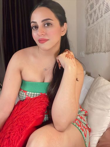 Woman in a colorful strapless dress with henna on her hand, sitting on a bed.