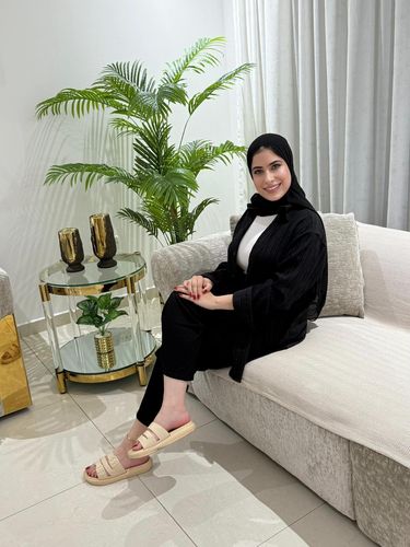 Young woman in black oversized shirt and beige slide sandals sitting on a couch.
