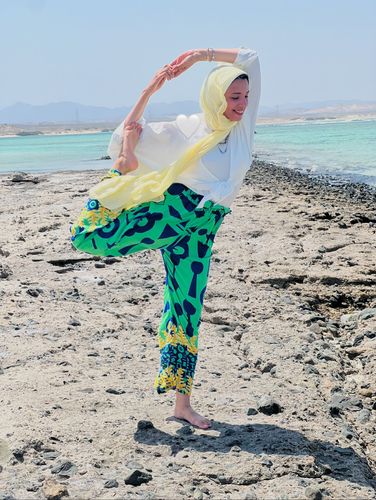Woman in vibrant patterned palazzo pants and hijab practicing yoga on a rocky beach